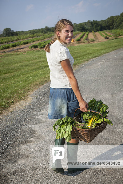 A girl in boots on a farm carrying a basket full of fresh produce.