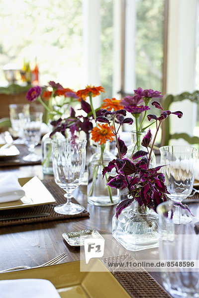 Ein für eine Mahlzeit gedeckter Tisch mit Gläsern und Glasvasen aus leuchtend bunten Orangen- und Kastanienblüten als Mittelpunkt.
