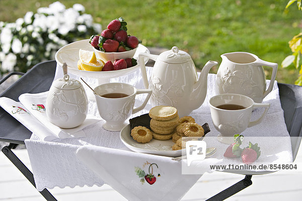 Ein traditioneller Nachmittagstee  der auf einem Teetablett in einem Garten mit weißem Porzellan und Erdbeeren serviert wird.