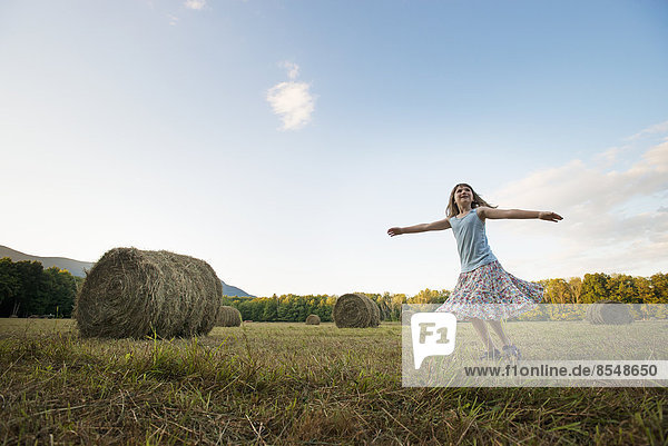 A field full of tall rounded hay bales  and a young girl dancing with her arms outstretched on the stubble field.