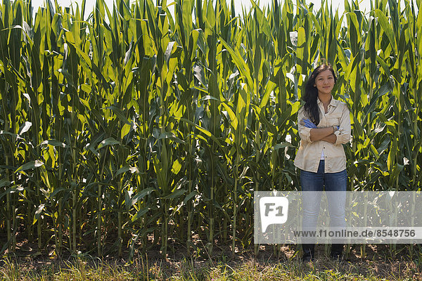 Eine junge Frau steht mit verschränkten Armen vor einer sehr hohen Maisernte auf dem Feld.