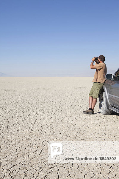 A man standing in a dry desert  looking through binoculars and leaning against a truck