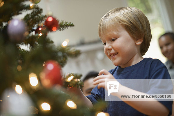 Ein kleiner Junge hält Weihnachtsschmuck in der Hand und legt ihn auf den Weihnachtsbaum.