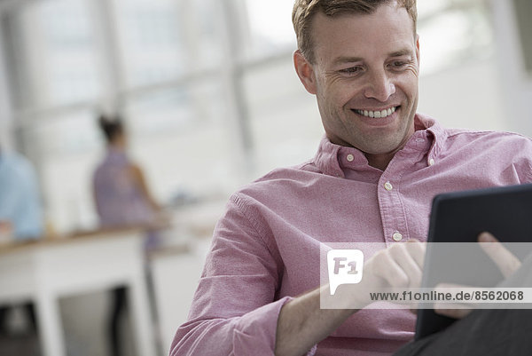 An open plan office in New York City. A man in a pink shirt sitting smiling  using a digital tablet.