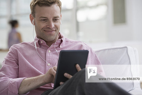 An open plan office in New York City. A man in a pink shirt sitting smiling  using a digital tablet. Wearing earphones.