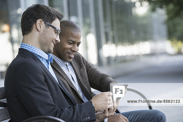 Geschäftsleute in der Stadt. Unterwegs in Kontakt bleiben. Zwei Männer sitzen auf einer Parkbank im Freien und schauen auf ein digitales Tablet.