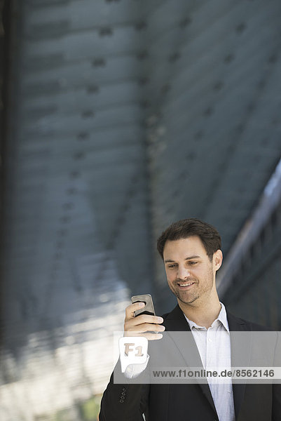 Geschäftsleute in der Stadt. Unterwegs in Kontakt bleiben. Ein Mann in schwarzer Jacke und Hemd mit offenem Kragen  der ein Smartphone benutzt.