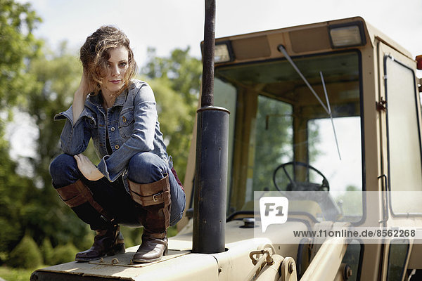 Eine junge Frau in Jeansjacke und Stiefeln auf der Motorhaube eines Traktors.