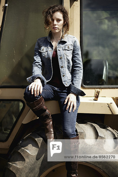 A young woman in denim jacket and boots on the hood of a tractor.