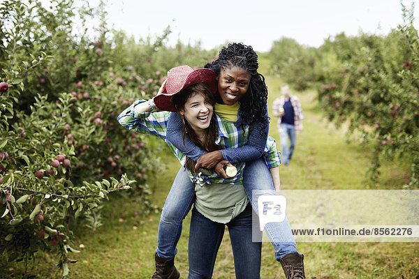 Rows of fruit trees in an organic orchard. A young woman giving another a piggyback.