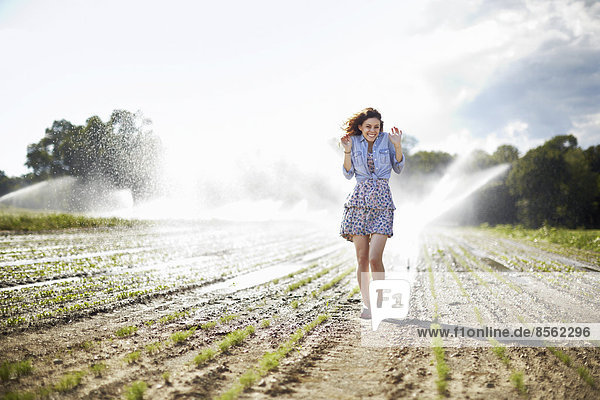 A young woman in denim jacket standing in a field  irrigation sprinklers working in the background.