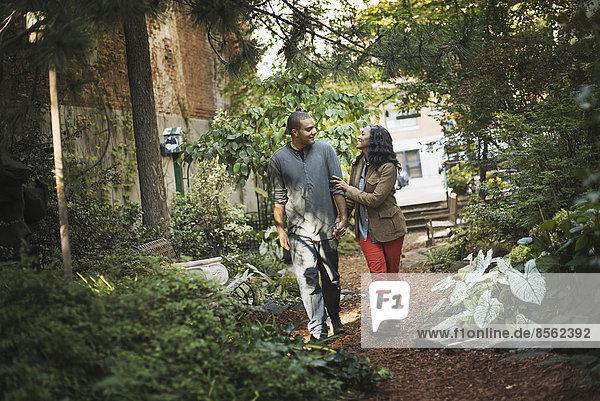 Szenen aus dem städtischen Leben in New York City. Ein Mann und eine Frau  ein Paar beim Spaziergang durch den Park  durch einen begrünten Raum mit Bäumen und grünem Laub und einer Bank.