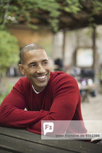 Scenes from urban life in New York City. A man in a red jumper seated at a bench  smiling.