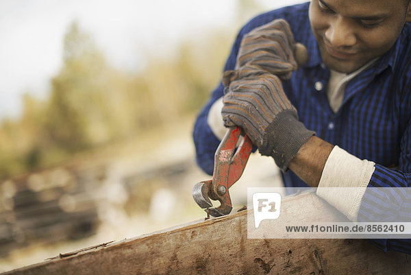 Ein Mann arbeitet auf einem ehemaligen Holzlager. Er benutzt ein Werkzeug  um Metalle aus einem Stück Altholz zu entfernen.