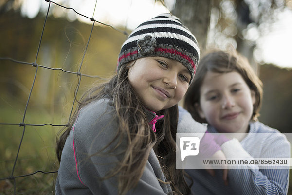Two young girls side by side outdoors on a farm. Leaning on a fence post.