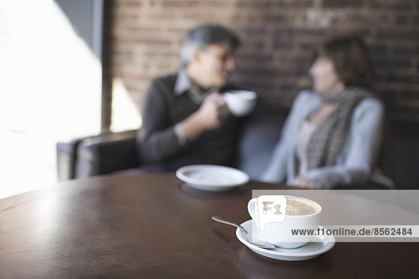 Zwei Personen sitzen in einem Café. Ein Mann hält ein weißes Porzellan in der Hand und trinkt. Sitzt neben einer Frau. Ein Tisch mit einer großen vollen Tasse Cappuccino-Kaffee.