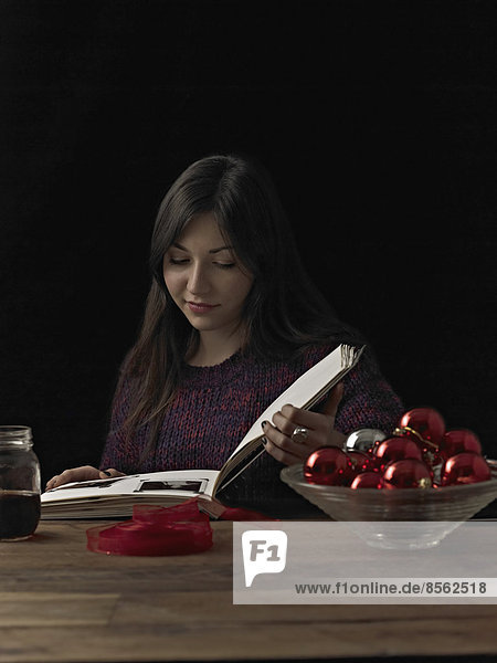 Eine Frau  die an einem Tisch sitzt und ein Buch liest. Eine Glasschale mit rot glänzenden Kugeln und eine Spule mit rotem Band.