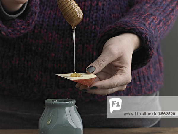 Eine Frau  die Honig von einem runden hölzernen Nieselstab auf eine Apfelscheibe träufelt.