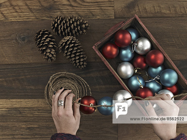 A woman threading Christmas shiny round ornaments on a piece of string. A small group of pine cones.