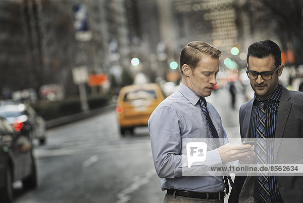 Two men standing together looking at a cell phone display on a busy street at dusk.