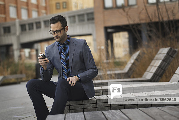 A man in a formal jacket and tie  sitting on a bench outside a city building  checking his phone for messages.
