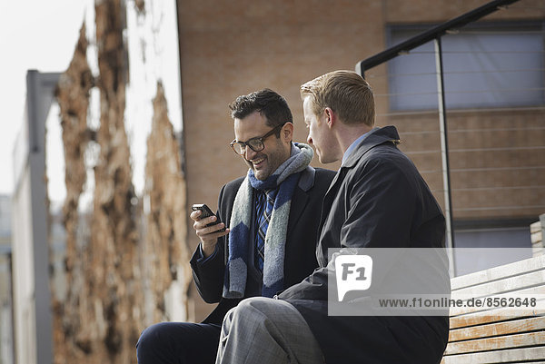 Zwei Männer sitzen auf einer Bank vor einem großen Gebäude in der Stadt. Einer überprüft sein Mobiltelefon.