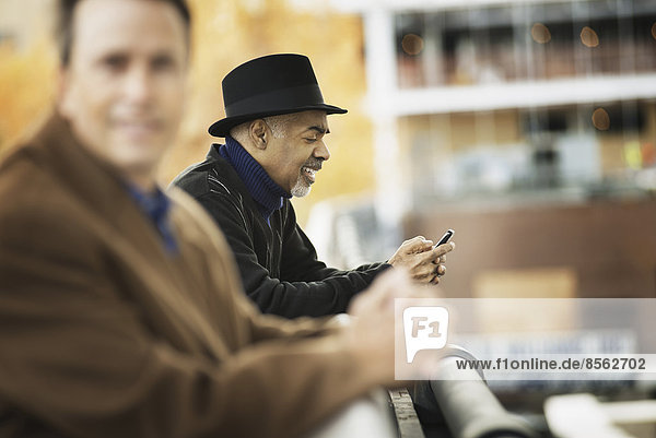 Stadtleben. Berufstätige Menschen unterwegs  in Kontakt bleiben  Mobiltelefone benutzen. Zwei Männer in Mänteln  die sich an ein Geländer lehnen.