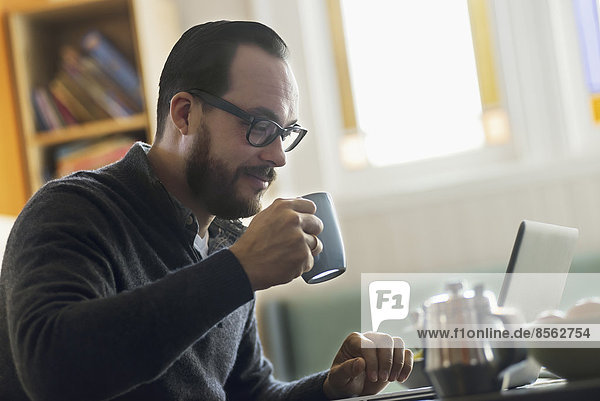 A bearded man having a drink of coffee. An open laptop computer on the counter.