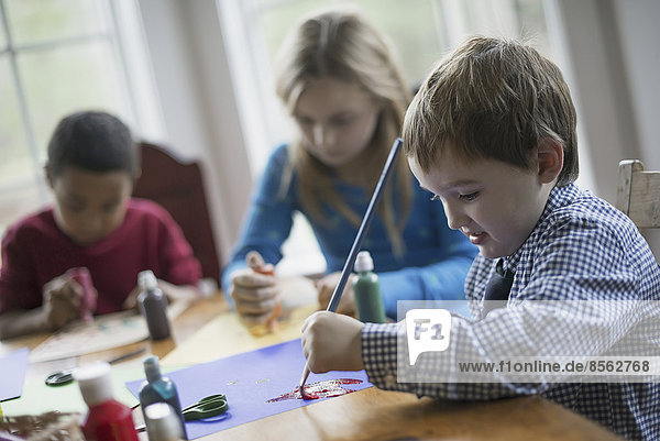 Kinder in einem Familienhaus. Drei Kinder sitzen an einem Tisch und verwenden Leim und Farbe  um Dekorationen zu schaffen.