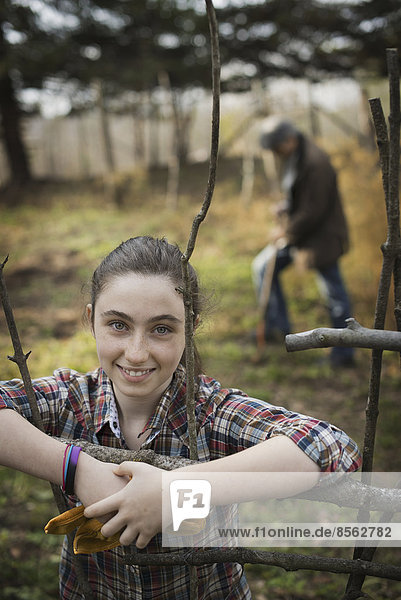 Ein junges Mädchen lehnt an einem Zaun  auf einem Biohof. Im Hintergrund ein Mann  der den Boden umgräbt.