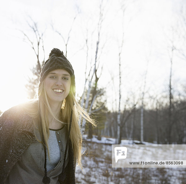 Eine junge Frau mit blonden Haaren und einer gestrickten Wollmütze und einem Wollmantel. Draußen an einem Wintertag. Schnee auf dem Boden.