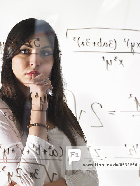 Eine junge Frau betrachtet eine mathematische Gleichung  die mit einem schwarzen Marker auf eine durchsichtige  durchsichtige Wand geschrieben wurde.