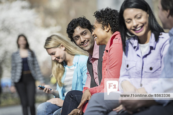 Menschen im Frühling in der Stadt im Freien. Fünf Menschen sitzen in einer Reihe  einer schaut auf ein Mobiltelefon. Eine Frau nähert sich in der Ferne.