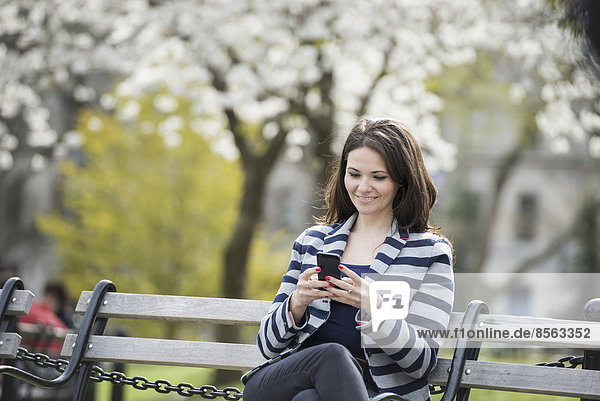 Draußen in der Stadt im Frühling. Park von New York City. Weiße Blüte an den Bäumen. Eine Frau sitzt auf einer Bank und hält ihr Mobiltelefon.