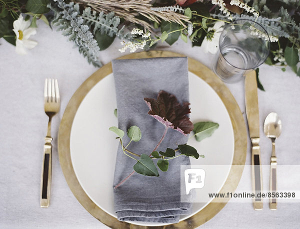 A table top covered with a white cloth. A place setting with cutlery and plate. A napkin and a foliage table decoration.