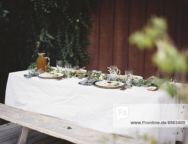 A table laid for a special meal. Place settings  with plates and cutlery. Glasses. A white table cloth and bench seat. Plates of food.