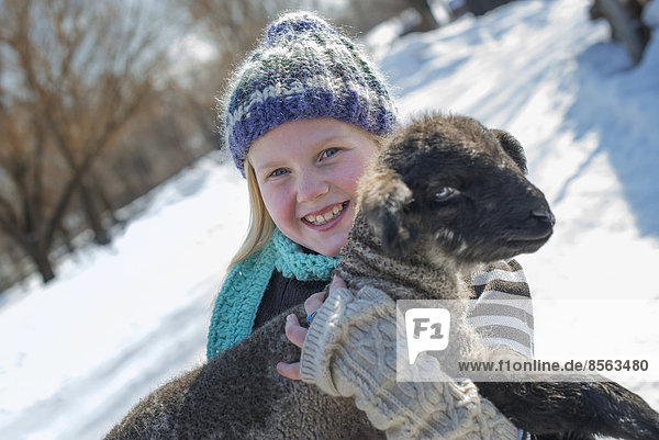 Winterlandschaft mit Schnee auf dem Boden. Ein junges Mädchen hält ein junges Lamm.