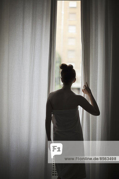 A young woman with her hair up  wearing a bath towel  looking through long curtains at a window.