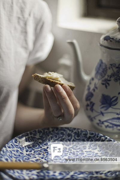 Ein Frühstückstisch mit einer hohen Kaffeekanne und einem blau-weißen Porzellanteller. Eine Frau sitzt mit einem Stück Butterbrot in der Hand.