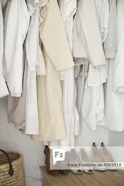 Ein Schrank mit cremefarbenen und weißen Kleidern  Jacken  Hemden und Tuniken  die aufgehängt sind. Eine Reihe Damenschuhe  die ordentlich auf dem Boden angeordnet sind.