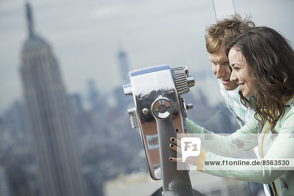 New York City. Eine Aussichtsplattform mit Blick auf das Empire State Building. Ein junges Paar schaut durch das Teleskop.