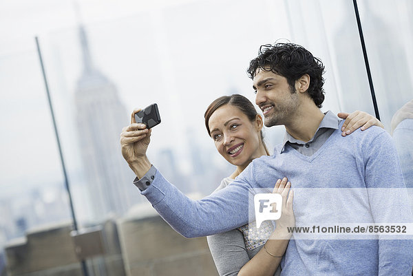 New York City. Eine Aussichtsplattform mit Blick auf das Empire State Building. Ein junges Paar  das mit einem Mobiltelefon fotografiert.