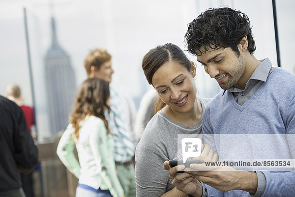 New York City. Eine Aussichtsplattform mit Blick auf das Empire State Building. Ein junges Paar  das mit einem Mobiltelefon fotografiert.