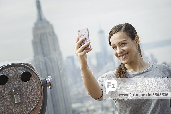 New York City. Eine Aussichtsplattform mit Blick auf das Empire State Building. Eine Frau  die mit ihrem Smartphone ein Foto von sich und dem Blick über die Stadt macht.