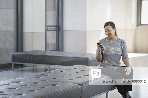 Urbaner Lebensstil. Eine junge Frau sitzt auf einem Sitz in einem Gebäude und benutzt ihr Mobiltelefon.