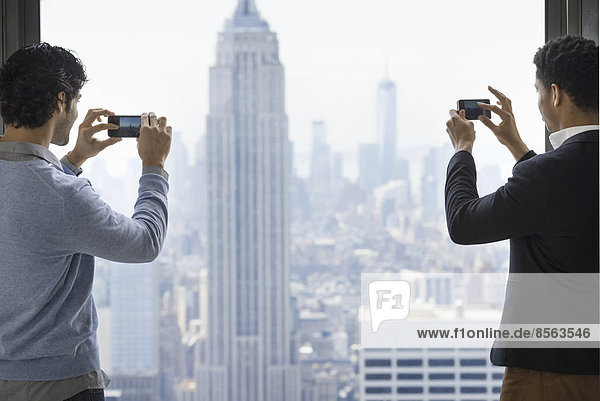Urbaner Lebensstil. Zwei junge Männer  die von einer Aussichtsplattform mit Blick auf das Empire State Building mit ihrem Telefon Bilder der Stadt aufnehmen.
