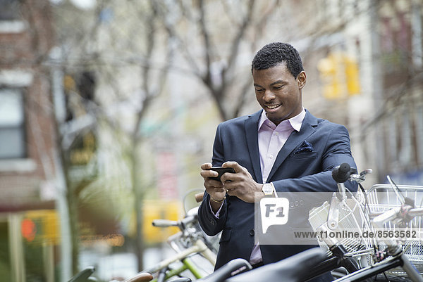 Stadtleben im Frühling. Ein junger Mann in einem blauen Anzug  an einem Fahrradparkplatz. Er überprüft sein Smartphone auf Nachrichten.