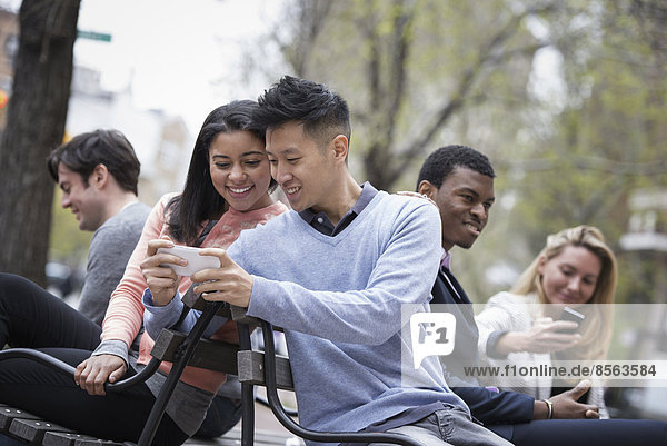 Stadtleben im Frühling. Jugendliche im Freien in einem Stadtpark. Sitzen auf einer Bank. Ein Mann zeigt einer Frau den Bildschirm seines Telefons  und drei weitere Personen überprüfen ihre Telefone.