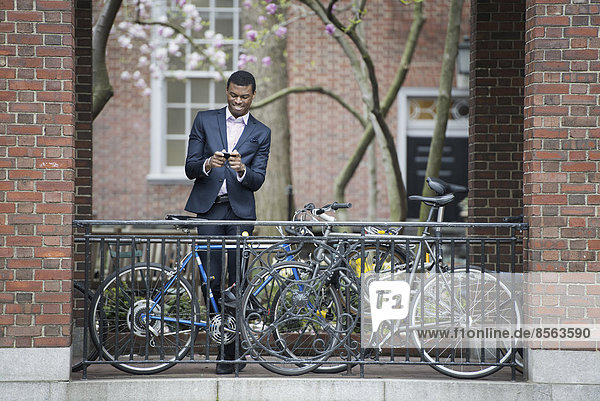 Stadtleben im Frühling. Jugendliche im Freien in einem Stadtpark. Ein Mann in einem Anzug  neben einem Fahrradparkplatz auf einem Bürgersteig. Er benutzt sein Smartphone.