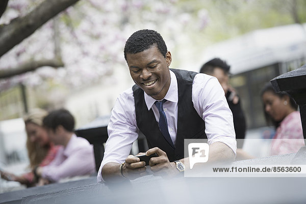 Stadtleben im Frühling. Jugendliche im Freien in einem Stadtpark. Ein Mann sitzt und schaut auf sein Smartphone  mit vier Personen in der Gruppe hinter ihm.